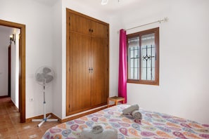 A bedroom with a bed, a wooden wardrobe, a standing fan, a window with a curtain, and a small stool.