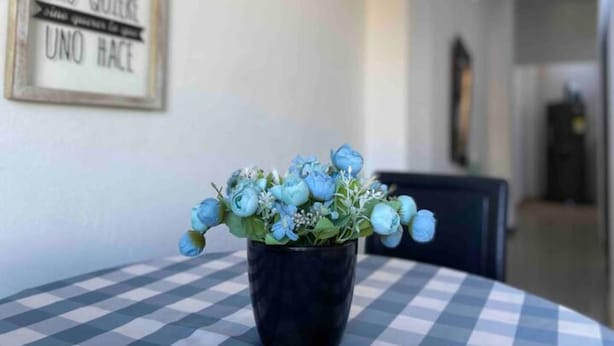 Una mesa con mantel a cuadros, un jarrón negro con flores azules y un cartel enmarcado en la pared.