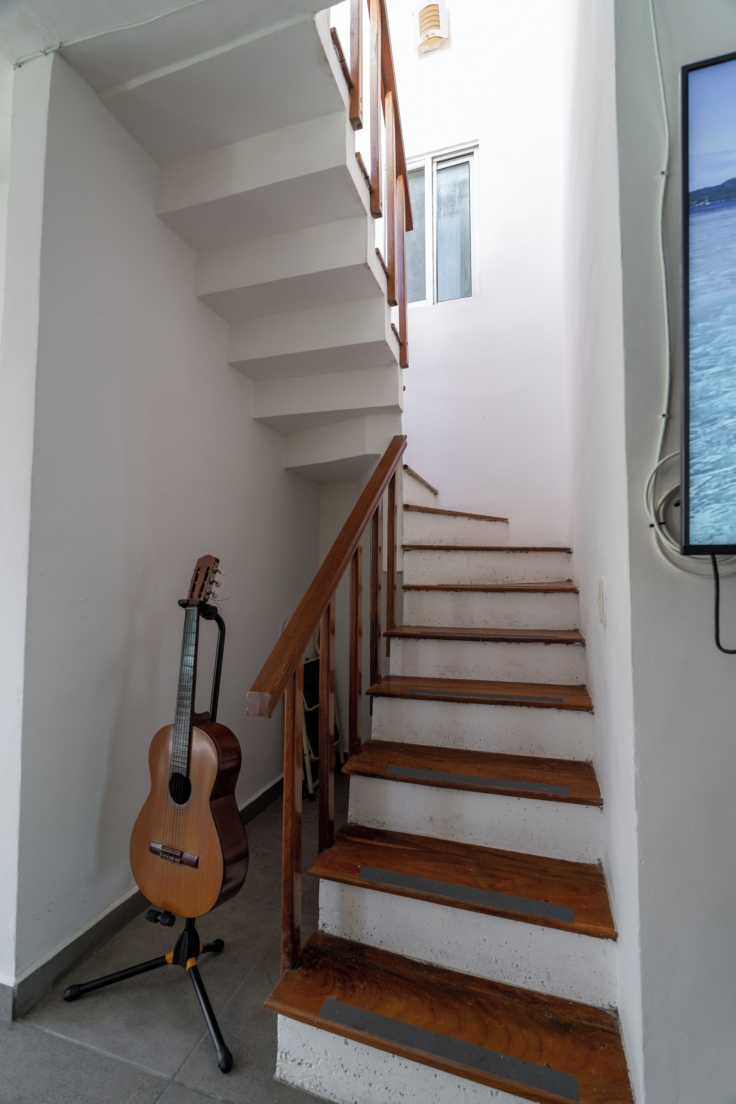 Un escalier avec des marches en bois et une rampe, une guitare appuyée contre le mur et une fenêtre donnant sur la mer.