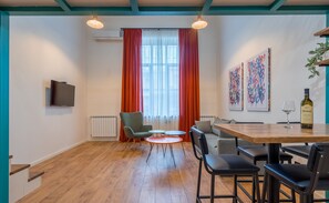 Een moderne woonkamer met houten vloer, een flatscreen televisie, een eettafel met stoelen, een bank en een raam met rode gordijnen.