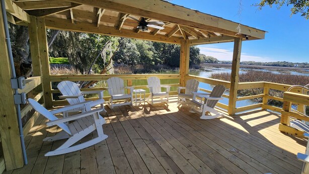 A wooden deck with white Adirondack chairs and a covered seating area overlooking a body of water and marshland.