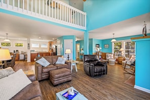 A spacious living area with a high ceiling, a kitchen in the background, and a view of the outdoors through large windows.