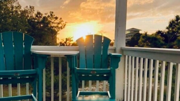 A balcony with two green Adirondack chairs and a white railing, overlooking a sunset.