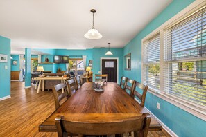 A dining area with a wooden table and chairs, a ceiling light, and a view through a window with blinds.