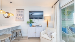 Una habitación con pared blanca, televisor montado, sofá, mueble pequeño y ventana decorada con un ancla.