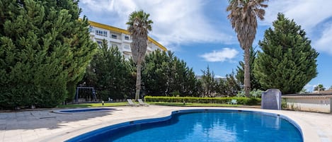 A swimming pool with lounge chairs, surrounded by trees and a building with a yellow roof.