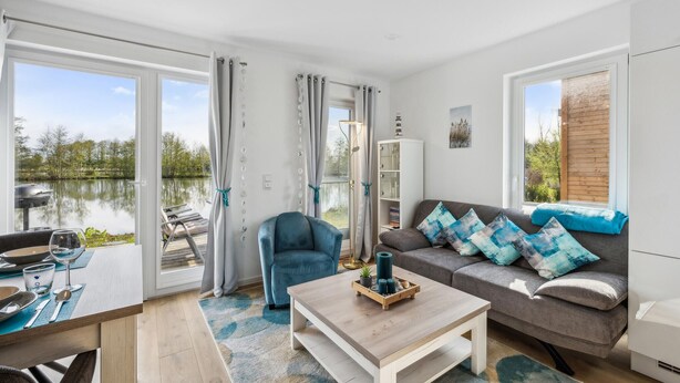 A modern living room with a view of a lake, a dining area with outdoor seating, and a cozy seating area with a sofa and cushions.