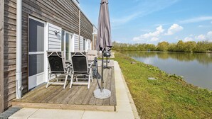 A wooden deck with outdoor furniture, a grill, and an umbrella by a house with a view of a lake.