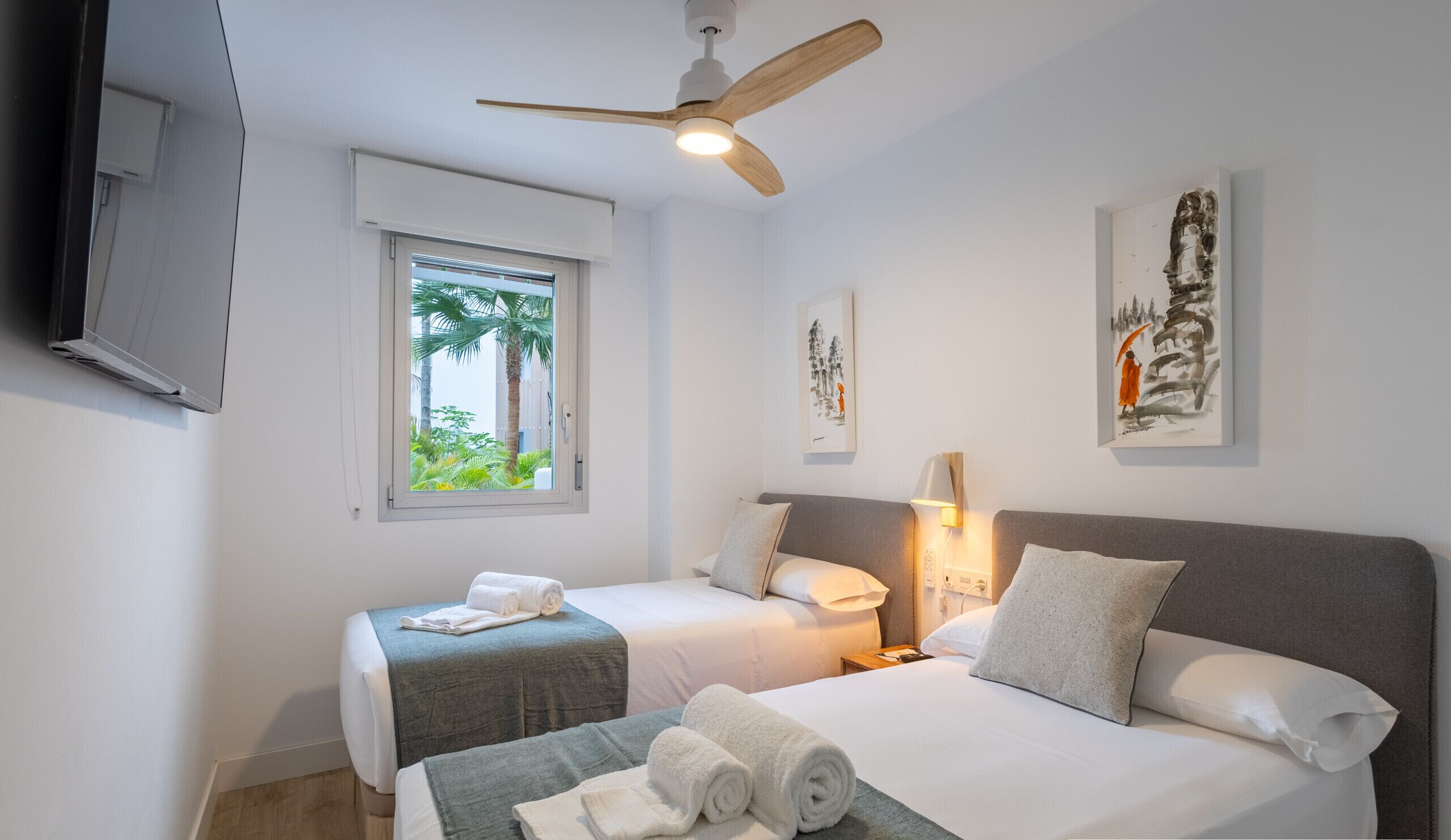 A hotel room with two beds, a ceiling fan, a TV, a window with a view of palm trees, and artwork on the walls.