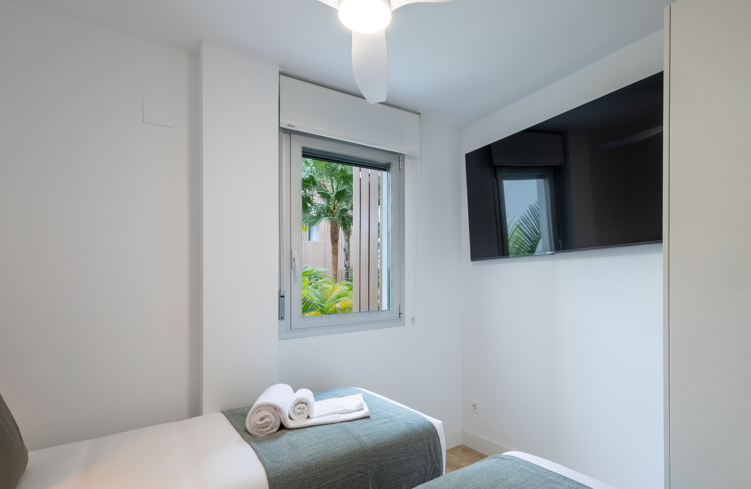Una habitació petita i moderna amb un llit individual, una finestra amb vista a palmeres i un televisor de pantalla plana muntat a la paret.