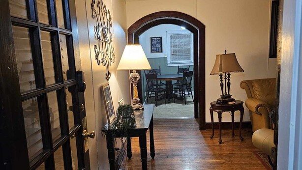 Un pasillo con suelo de madera, una mesa con sillas, una lámpara y una puerta con paneles de vidrio.