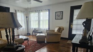 Un salón con sofá, sillón y reposapiés, una mesita de centro, una lámpara y una lámpara de pie. Se ve una ventana con cortinas y un cuadro enmarcado en la pared.