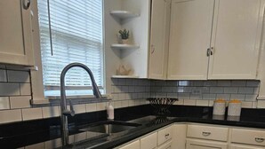 Una cocina con armarios blancos, encimera negra y ventana con persianas.