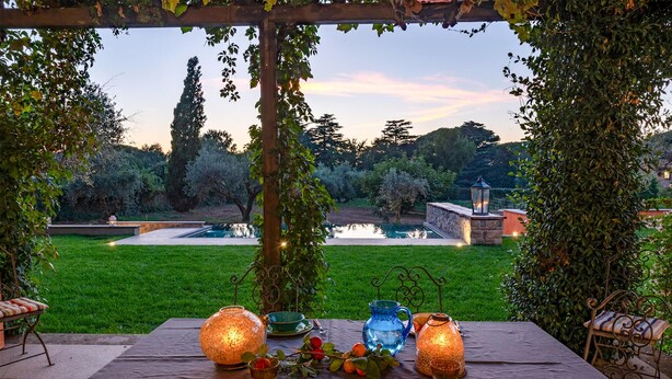 An outdoor dining area with a table set for two, surrounded by greenery and a view of a garden and a pond. - Lovely home in Rocca di Papa (Rocca di Papa)
