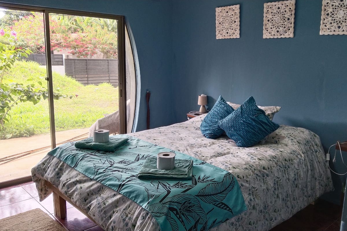 A bedroom with a bed, a window, a wall with decorative panels, and a view of greenery outside.