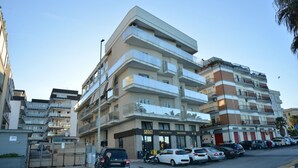 Un edificio residenziale a più piani con balconi, un ingresso con cancello e auto parcheggiate sulla strada.