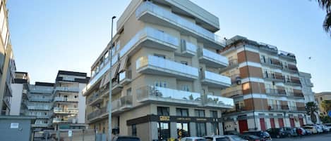 A multi-story residential building with balconies, a gated entrance, and parked cars on the street.