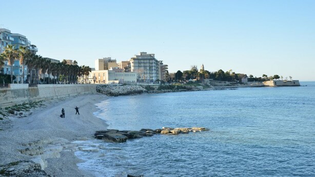 Una spiaggia con edifici sullo sfondo, un litorale roccioso e un cielo sereno.