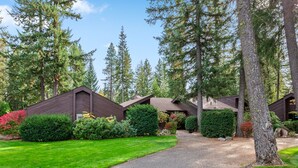 A row of cabins with a gravel driveway leading to a wooden gate, surrounded by dense forest and well-maintained lawns. - Twin Lakes Village Home—Golf Retreat, Community Pool, Corner Lot, and Single-Level Living! (Rathdrum)
