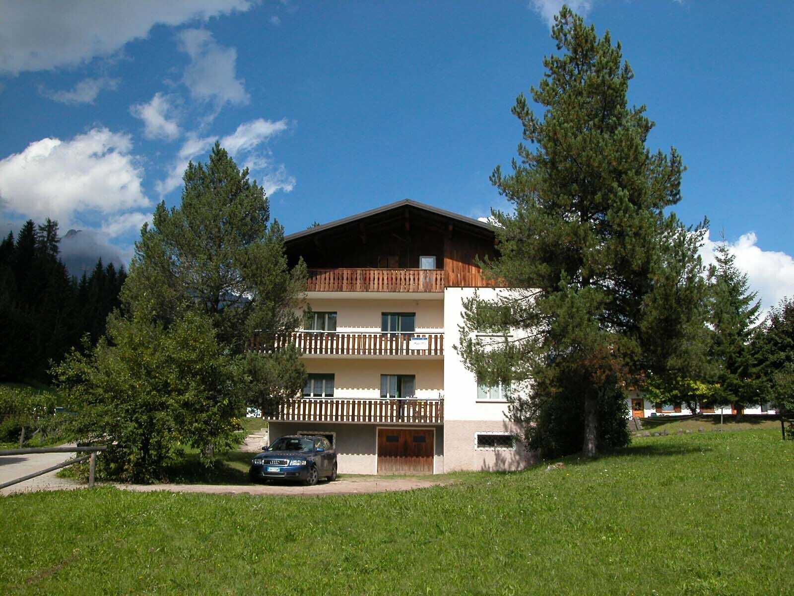 A multi-story building with a wooden balcony, surrounded by trees and a clear sky.