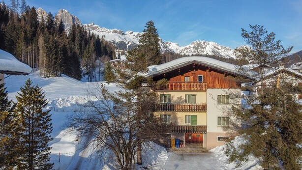 A two-story chalet with a wooden balcony, surrounded by snow-covered mountains and pine trees. - Casa Iris 2 (Falcade)