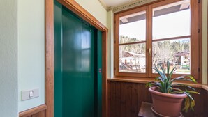 Une pièce avec une porte verte, une fenêtre donnant sur une maison et des arbres, et une plante en pot posée sur une table en bois.