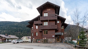 A multi-story wooden building with balconies, a stone foundation, and a parking area in front.