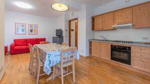 A compact living space with a dining area, a red sofa, wooden cabinets, and a kitchenette. - Villa Licia trilo (Falcade)