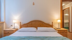 A neatly made bed with white linens and a blue-striped duvet, flanked by wooden nightstands with lamps, in a room with a wooden headboard and a small wall decoration. - Villa Licia trilo (Falcade)