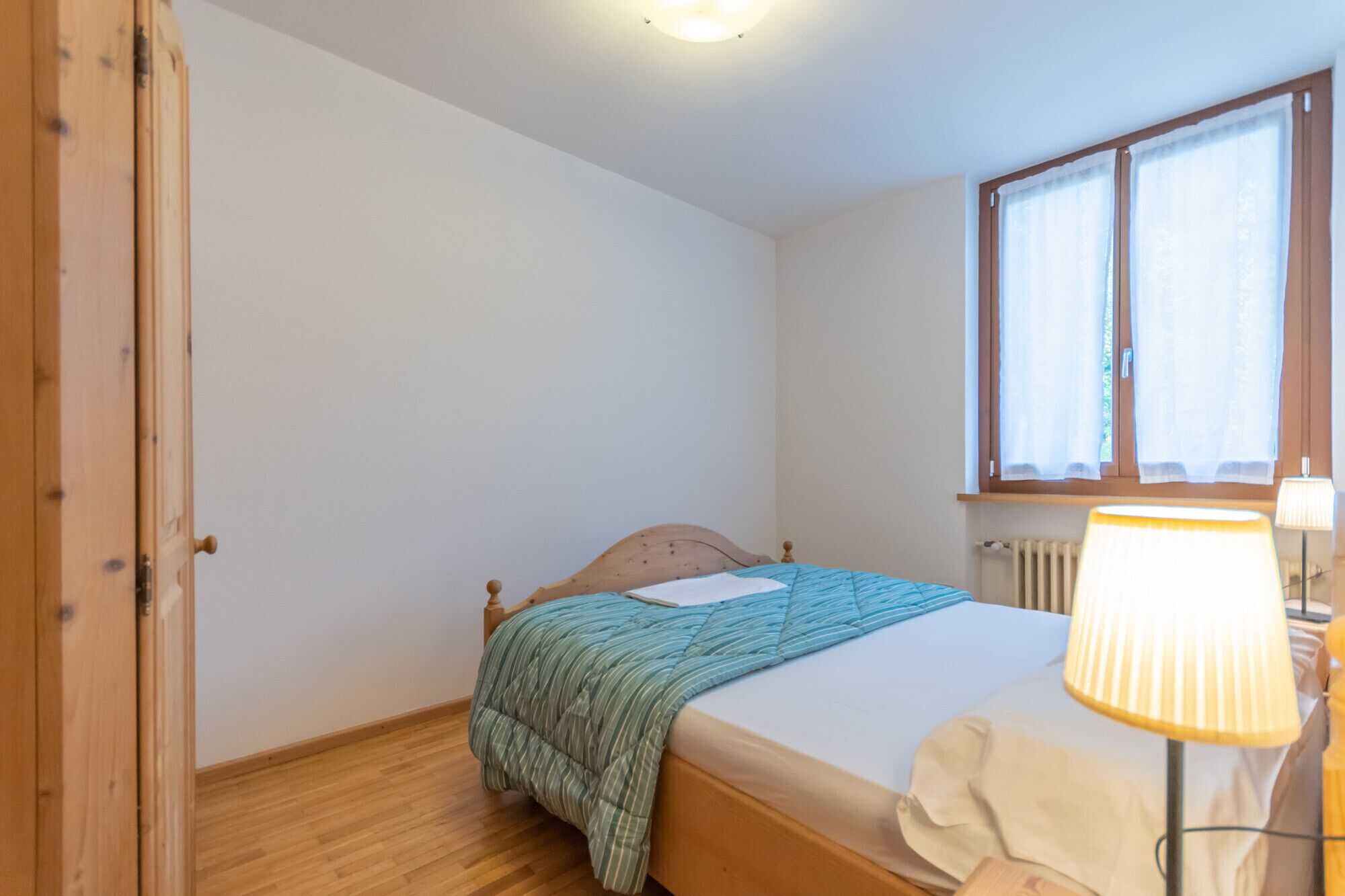 Un dormitorio con cama de madera, una ventana con cortinas, una lámpara de noche y un radiador.