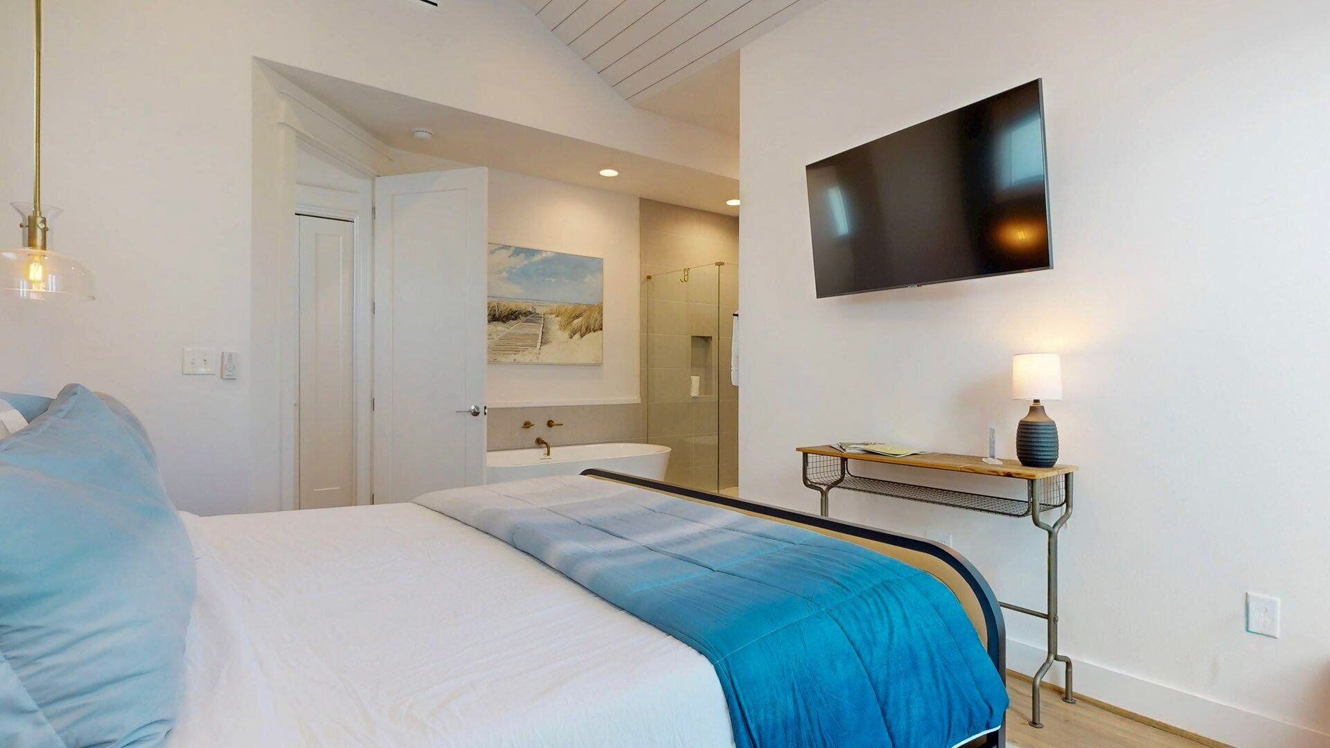 A modern bedroom with a large bed, a mounted TV, a bedside table with a lamp, and a bathroom visible through an open door.