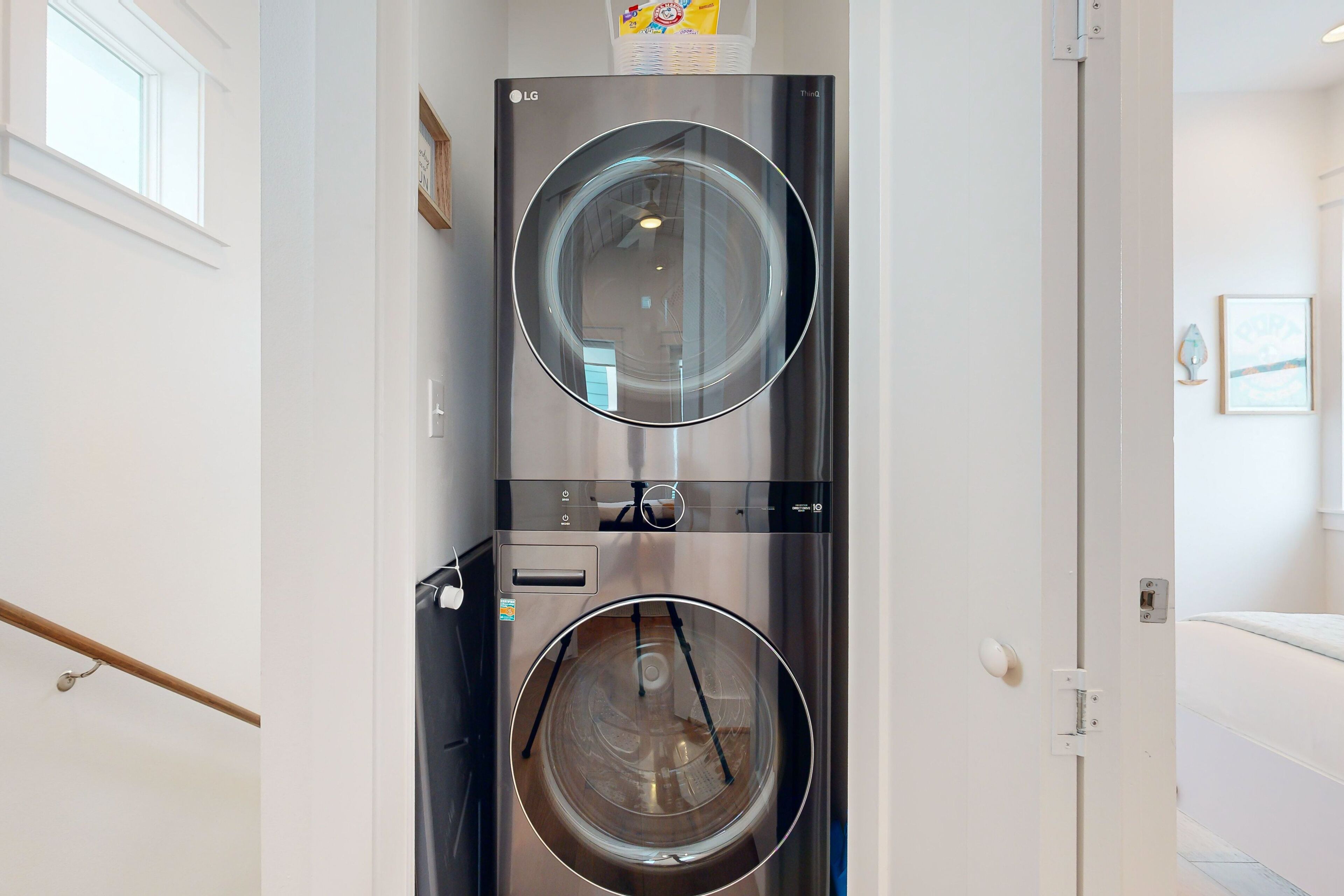 A modern, stainless steel washer and dryer unit in a clean, white-walled laundry room with a wooden handrail and a visible door.