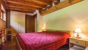 A bedroom with a wooden ceiling, a bed with a red quilt, a nightstand with a lamp, and a wooden wardrobe. - Casa Falcade 10 (Falcade)