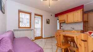 A bedroom with a bed, a wooden dining table with chairs, a kitchen area with wooden cabinets, and a window with a curtain.