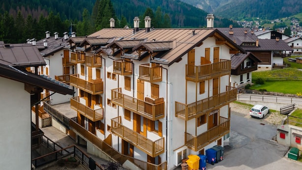 A multi-story building with balconies, a parking area, and a view of mountains in the background. - Casa Stella 8 (Falcade)