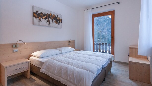 A bedroom with a wooden headboard, a bed with white bedding, a nightstand, a bedside table, a wall-mounted lamp, and a window with a view of trees. - Casa Zulian (Falcade)