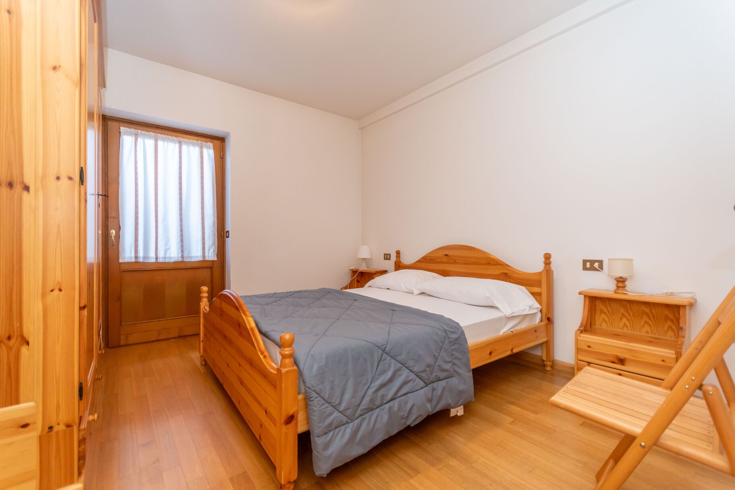 Een slaapkamer met een houten bed, nachtkastje, stoel en raam met gordijnen.