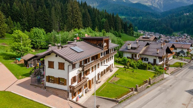 A traditional Alpine-style building with a balcony, surrounded by lush greenery and a mountainous backdrop. - Casa Morel (Falcade)