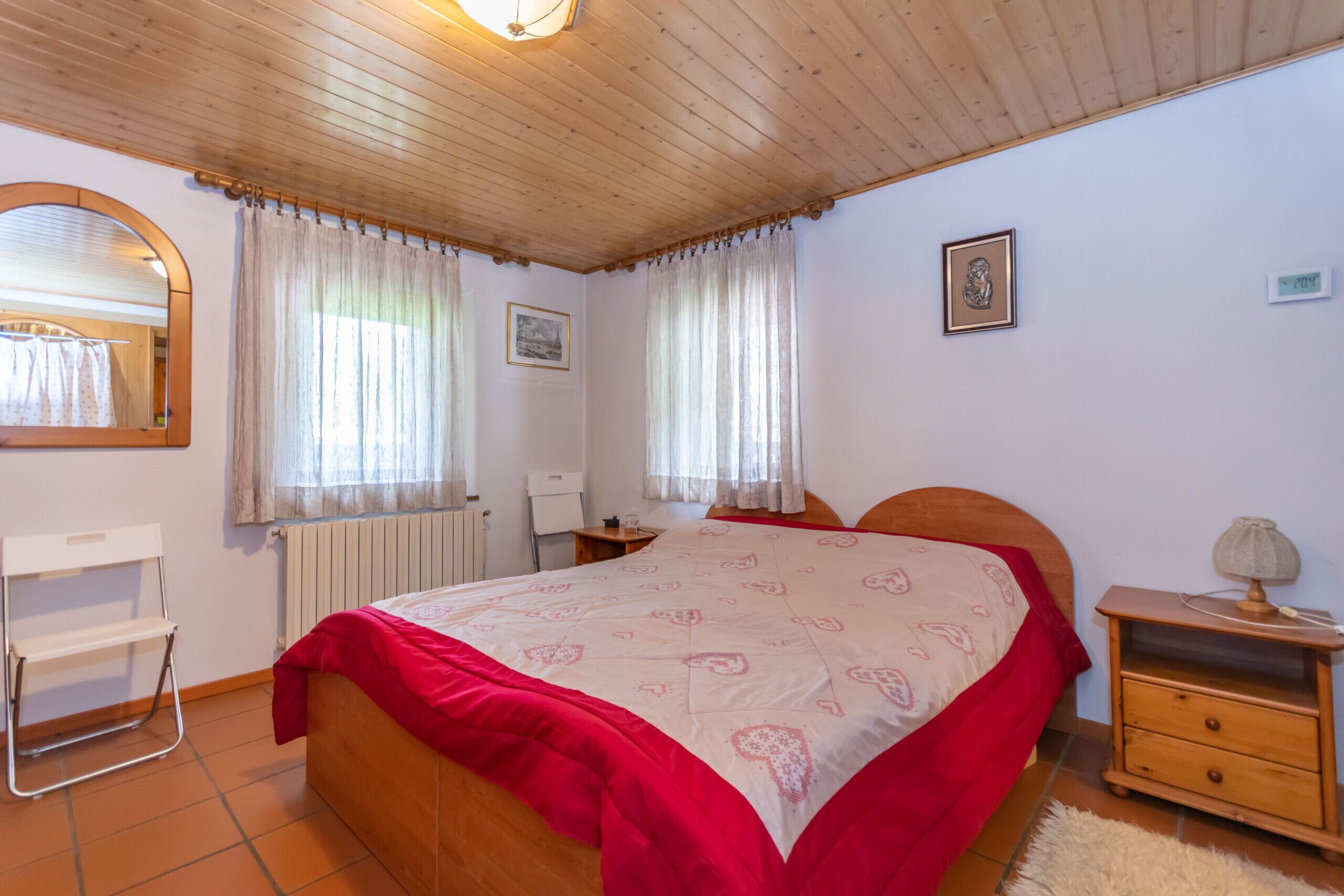 Una camera da letto con soffitto in legno, un letto con copripiumino rosso e bianco, un comodino con una lampada, una sedia e un termosifone.