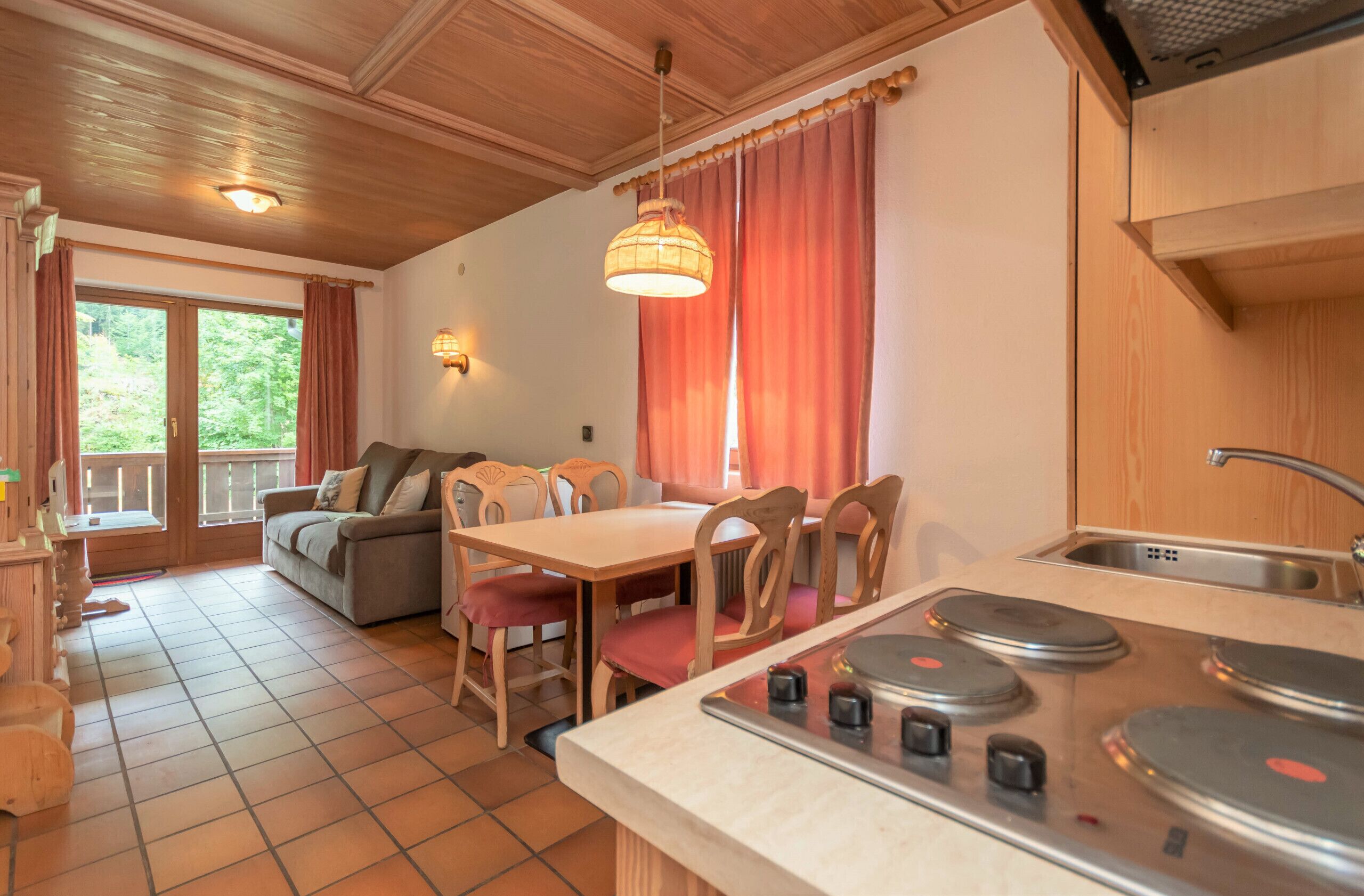 A cozy kitchen with wooden cabinets, a dining table, and chairs. There is a window with curtains and a view of greenery outside.