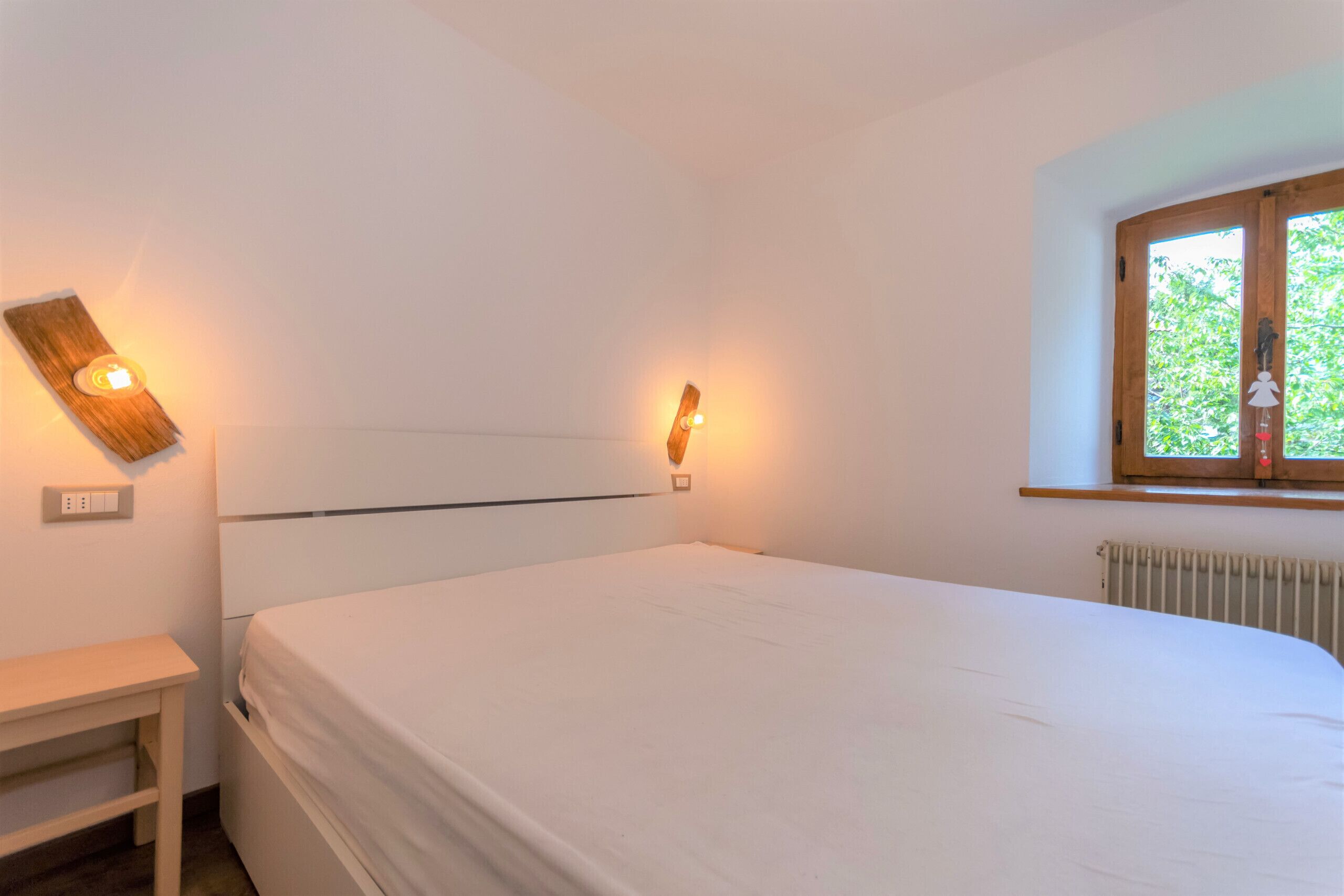Une chambre avec un grand lit, une table de chevet, une lampe fixée au mur et une fenêtre donnant sur un paysage verdoyant.