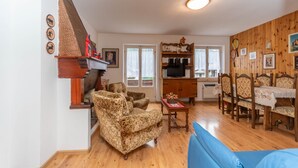 A living room with a sofa, armchairs, a wooden dining table with chairs, a fireplace, and a television. - Villa Elisena (Falcade)