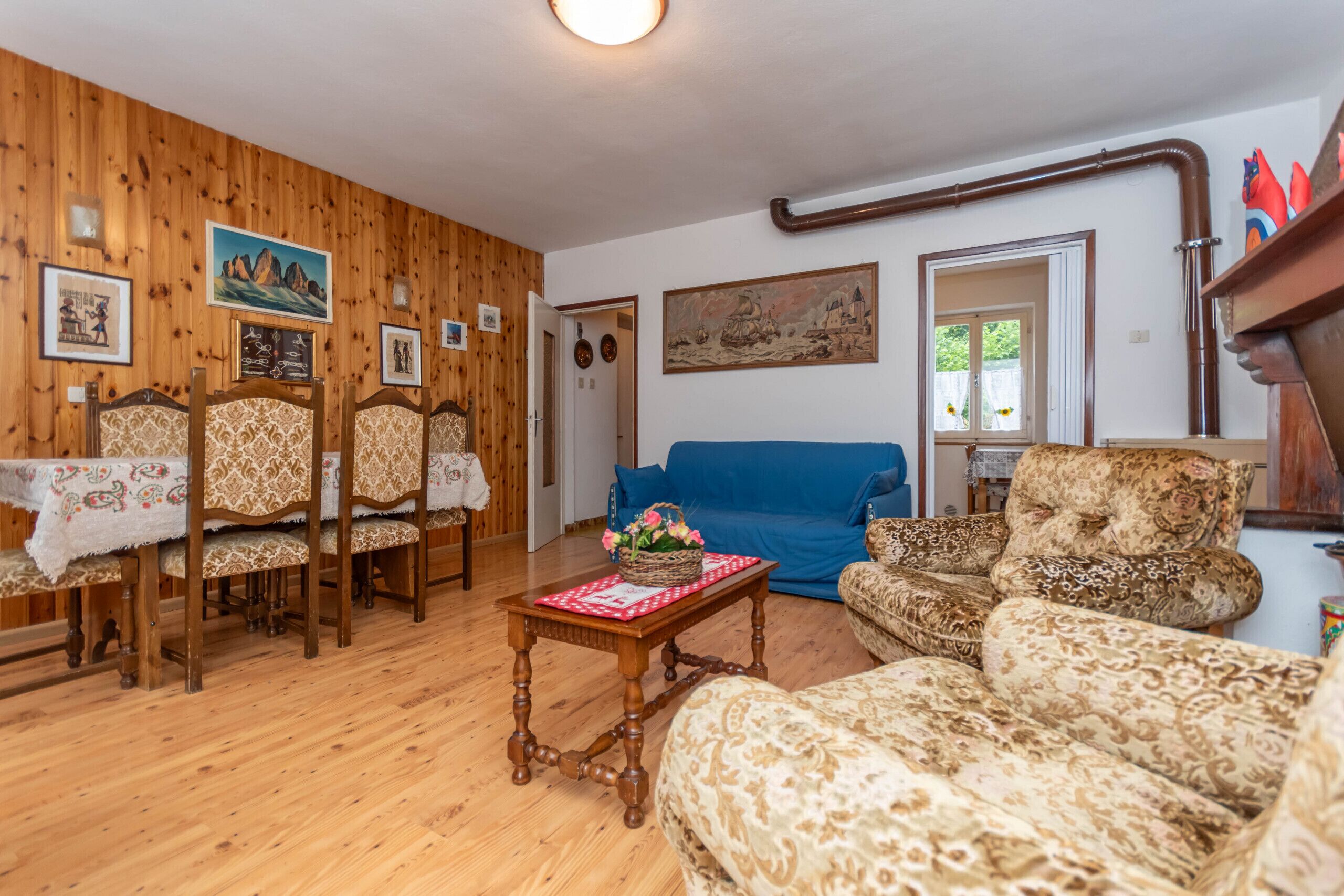 Una habitación con paneles de madera, un sofá azul, sillones estampados, una mesa de comedor con sillas y varias imágenes colgadas en las paredes.