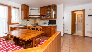 A kitchen with wooden cabinets, a microwave, and a dining area with a table and chairs. - Casa Alice (Falcade)