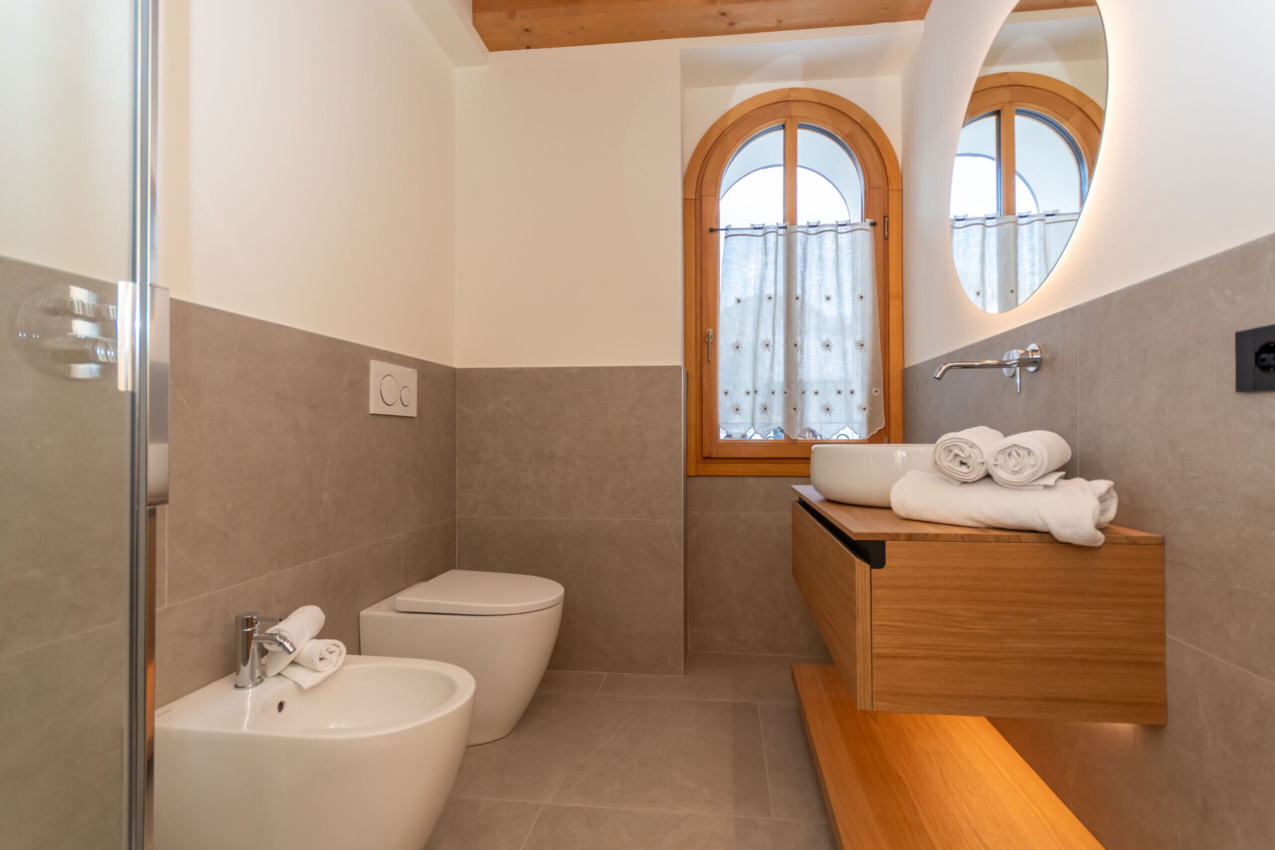 A modern bathroom with a toilet, sink, and vanity. There is a window with a curtain, and towels are neatly folded on the vanity.
