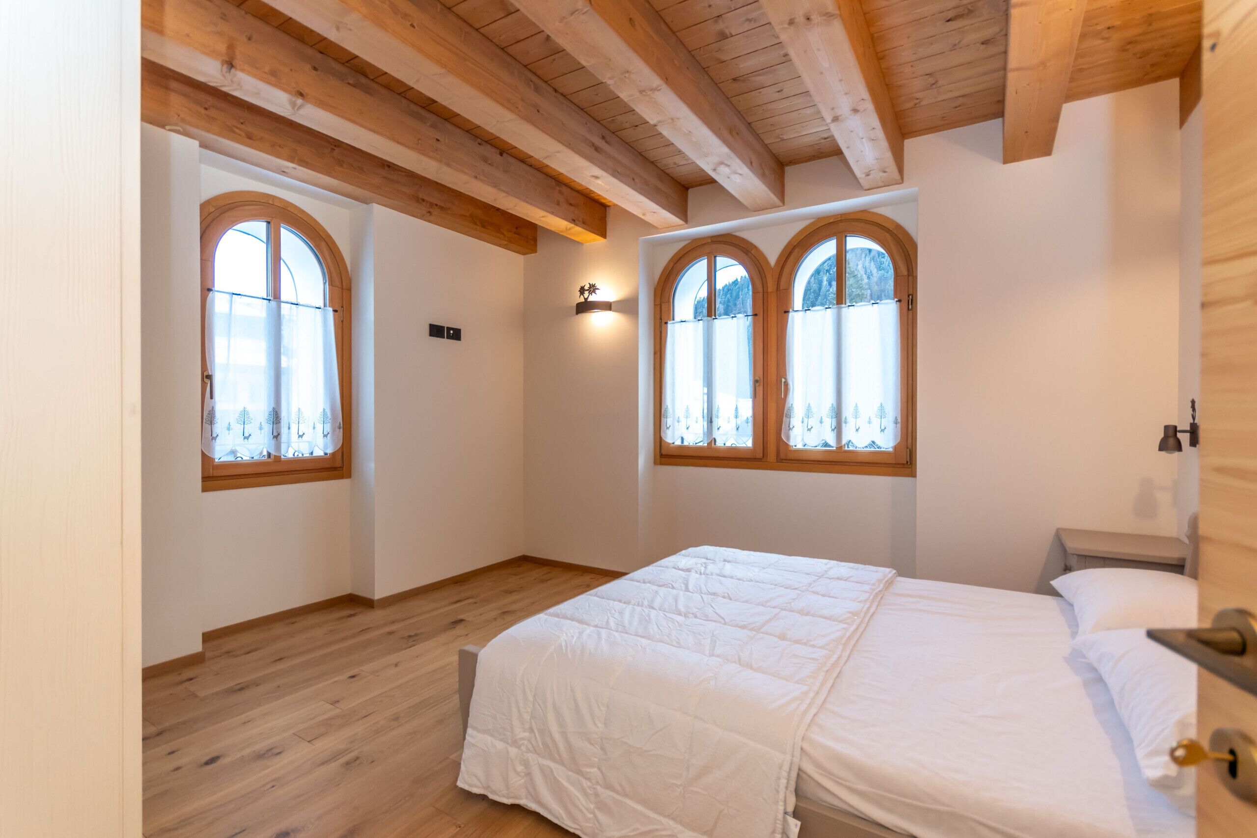 A bedroom with a wooden ceiling, two arched windows, a bed with white bedding, and a nightstand.