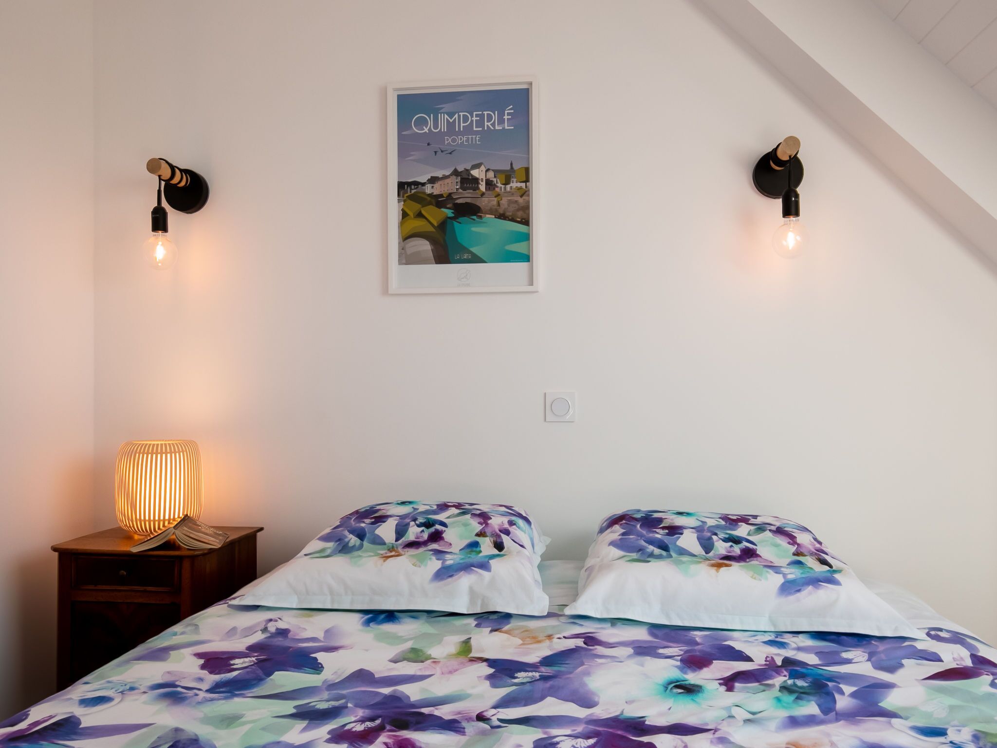 Une chambre à coucher avec un lit, une table de chevet, une lampe fixée au mur et un tableau encadré.