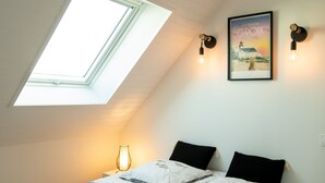 Une chambre avec une lucarne, un lit avec deux oreillers, une table de chevet avec une lampe et un tableau encadré au mur.