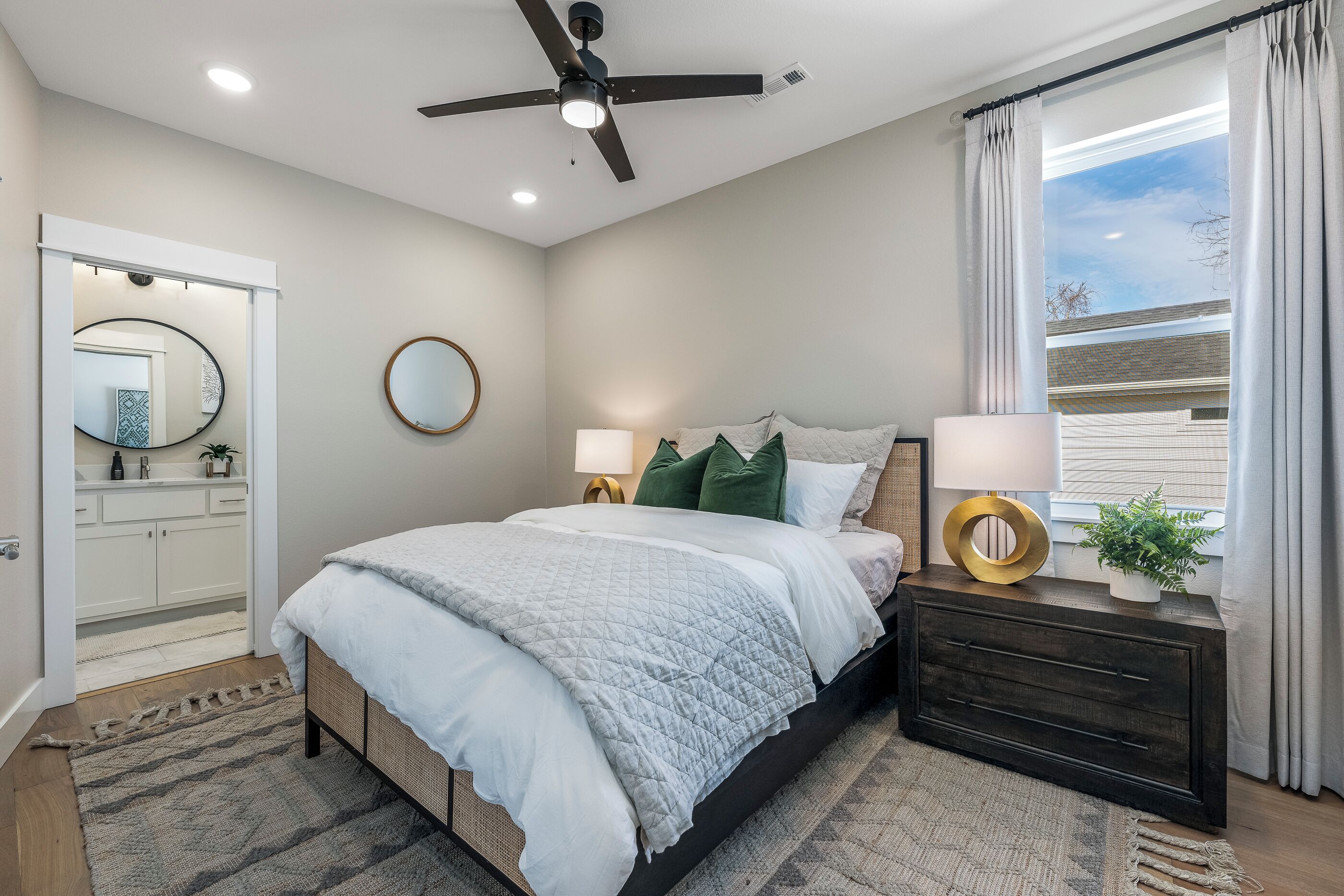 Quarto com cama, mesas de cabeceira, ventoinha no teto, janela com cortinas e vista para o exterior.