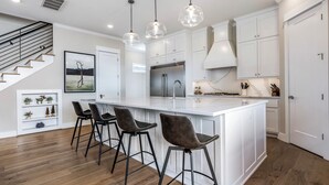A modern kitchen with a central island, bar stools, pendant lights, and a staircase. - New luxurious home - hot tub, firepit, & trails - Walk or ride to downtown! (Bentonville)